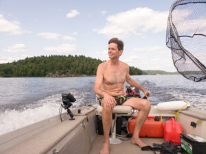 A man steers a small boat in a lake by the manual tiller on the motor. Shot from the front of the boat.