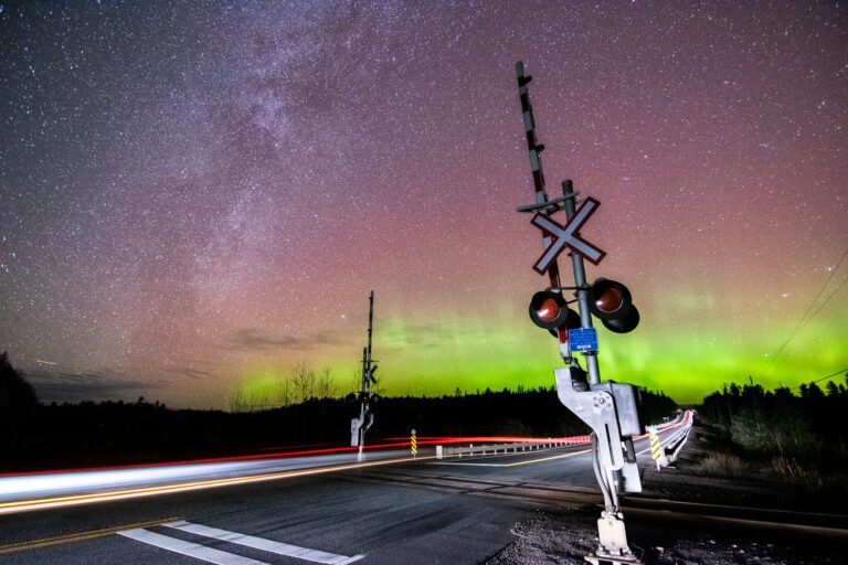 Northern lights and the milky way light the sky with a car's light streaks on a road and a railway crossing in the foreground.