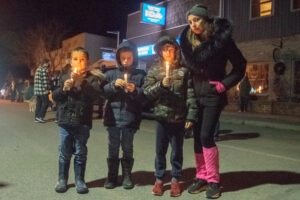A mother and three young boys holding candles stand on a street during a nighttime vigil.