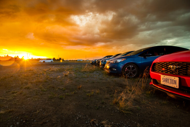 A bright sunset with dramatic clouds and a line of cars in the foreground, perpendicular to the camera.