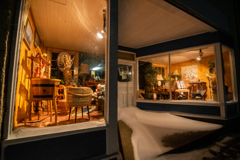 An odds-and-ends shop window bathed in warm light during a dark snowy night with a drift piling next to the front door.