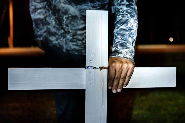 A man stands behind a white cross with his hand on it at night.